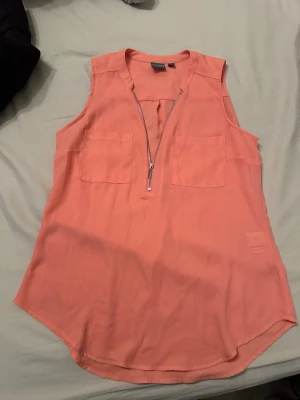 Korallfärgad ärmlös topp med dragkedja - Snygg ärmlös topp från Express i en fräsch korallfärg. Toppen har två bröstfickor och en dekorativ dragkedja framtill som ger en cool detalj. Perfekt till jeans eller kjol för en trendig look.