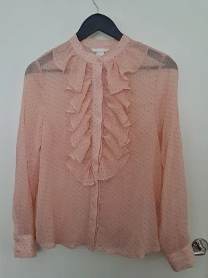 Ljusrosa blus - Ljusrosa blus med små svarta prickar och volangdetaljer framtill. Lätt genomskinlig och är mer åt ljusrosa/laxrosa i färgen str 36. Hittar inga defekter 🩷