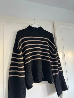 Svart stickad tröja med beige ränder - Säljer en oversized svart stickad tröja med beige  ränder. Tröjan har hög hals, långa ärmar och ribbstickade detaljer vid ärmslut och nederkant!!
