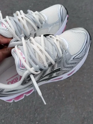 Vita och silver Asics Gel-Kayano 14 - Säljer ett par Asics Gel-Kayano 14 sneakers i vitt mesh med silverdetaljer och rosa inslag. Skorna har en chunky sula med rosa partier och klassisk Asics-logga på sidan. Perfekta för dig som gillar retrostil och sportig vibe.