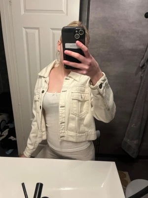 Vit cropped jeansjacka - Säljer en vit cropped jeansjacka med klassisk krage och silverfärgade knappar. Jackan har två bröstfickor med lock och knapp samt sömdetaljer framtill. Perfekt för dig som vill ha en stilren och trendig look.