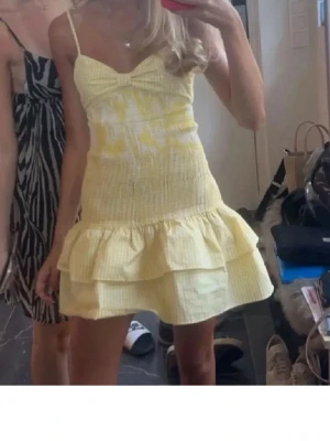 Gul volangklänning med smala axelband - Söt gul kortklänning med smala axelband och volangkant nedtill. Klänningen har en lätt rynkad struktur och en feminin passform med omlottliknande byst. Perfekt för varma dagar och sommarens alla tillfällen.