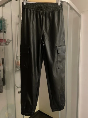 Svarta cargobyxor i fuskskinn Vero Moda - Snygga svarta cargobyxor från Vero Moda i fuskskinn. Byxorna har resår i midjan och vid bensluten samt stora cargofickor på sidorna. Perfekta för en trendig och edgy look. Materialet är mjukt och ger en cool läderkänsla.