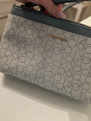 Calvin Klein axelväska med monogram - Snygg axelväska från Calvin Klein i beige och grått med klassiskt CK-monogram över hela väskan. Väskan har justerbar axelrem, guldfärgad logga och dragkedja upptill. Tillverkad i tåligt konstläder som ger en lyxig känsla.