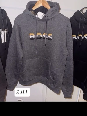 Grå BOSS hoodie med tryck - Mörkgrå hoodie från BOSS med stor logga i vitt, svart och gult på bröstet. Dom är helt nya materialet inuti är väldigt skönt och väldigt bra kvalité.