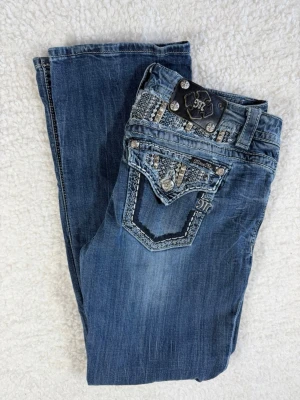 Blå Miss Me bootcut jeans W30 - Snygga blå bootcut jeans från Miss Me med dekorativa fickor och detaljer i silver på bakfickorna. Jeansen har slitningar och broderier som ger en cool look. Materialet är klassisk denim och passformen är bekväm med låg midja.