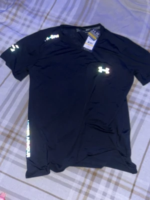 Svart tränings-tshirt från Under Armour - Snygg svart tränings-tshirt från Under Armour med reflekterande logga på bröstet och ärmen. Tillverkad i ett lätt och snabbtorkande material som passar perfekt till gymmet eller löprundan. V-ringad hals och diskret tryck längs sidan.