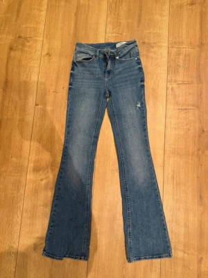 Blå bootcut jeans från Gina Tricot - Snygga blå bootcut jeans från Gina Tricot med klassisk femficksmodell och lätt slitning på låret. Jeansen har normal midja och är tillverkade i mjuk denim med lite stretch för skön passform. Perfekta för en trendig och avslappnad look.
