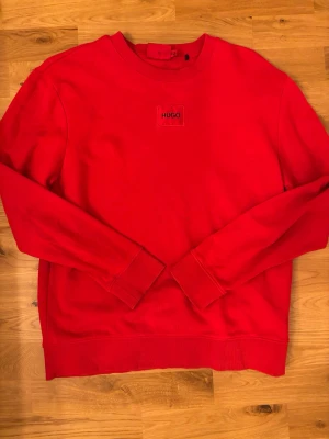 Röd sweatshirt från Hugo Boss  - Säljer en röd sweatshirt från Hugo med rund halsringning och diskret logga på bröstet. Tröjan har långa ärmar, ribbade muddar och är tillverkad i mjukt bomullsmaterial. Perfekt för en avslappnad och stilren look.