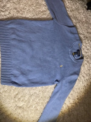 Ljusblå stickad tröja från Polo Ralph Lauren - Snygg ljusblå stickad tröja från Polo Ralph Lauren med klassisk gul broderad logga på bröstet. Tröjan har rund halsringning, långa ärmar och breda ribbade muddar vid ärmslut och nederkant. Perfekt för lager på lager och enkel att matcha. Jag köpte den från kidsbrandstore februari 2026 men den var för liten.