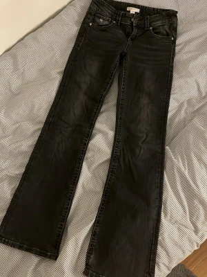 Svarta bootcut jeans med fickdetaljer - Snygga svarta bootcut jeans med slitningar och silverknappar på bakfickorna. Klassisk femficksmodell med låg midja och raka ben. Jeansen är tillverkade i ett mjukt denimtyg som ger en skön passform och cool look.