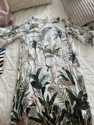 Vit blommig långklänning H&M - Säljer en vit långklänning från H&M med tropiskt blommönster och leoparddetaljer. Klänningen har lång ärm och är tillverkad i ett lätt, mjukt material som känns skönt mot huden. Perfekt för dig som gillar färgstarka prints och naturinspirerade plagg.