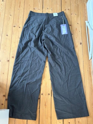 Grå kostymbyxor från Kings&queens - Snygga grå kostymbyxor från J.LUX med raka, vida ben,hög midja och klassiska passpoalfickor bak. Aldrig använda,säljs inte mer.