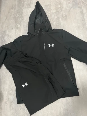 Under Armour - Säljer ett svart träningsset från Under Armour med jacka och byxor. Jackan har huva, dragkedja och reflekterande detaljer samt logotyp på bröstet. Byxorna har elastisk midja och logotyp på benet. Perfekt för löpning eller gym.
