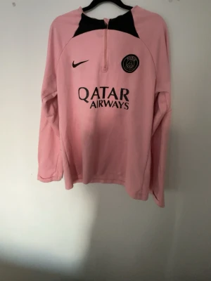 Rosa PSG träningströja från Nike - Säljer en långärmad rosa träningströja från PSG med svart Nike-logga och PSG-emblem på bröstet. Tröjan har halv dragkedja framtill och svart detalj vid halsen. Tillverkad i ett lätt och funktionellt material, perfekt för träning eller uppvärmning.