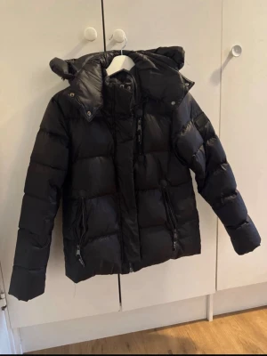 Svart pufferjacka från Amor - Snygg svart pufferjacka från Amor med huva och dragkedja framtill. Jackan har två stora fickor med dragkedja och en stilren patch på ärmen. Tillverkad i glansigt material som ger en modern look och håller dig varm under kalla dagar.Köp inte från hemsidan, tog tre månader att komma! Pris kan diskuteras 