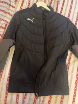 Svart vindjacka från Puma - Sportig svart vindjacka från Puma med hög krage och dragkedja framtill. Jackan har ett stilrent, quiltat mönster på bröstet och är tillverkad i lätt polyester. Perfekt för dig som gillar en clean look och vill ha en jacka som funkar till många outfits.
