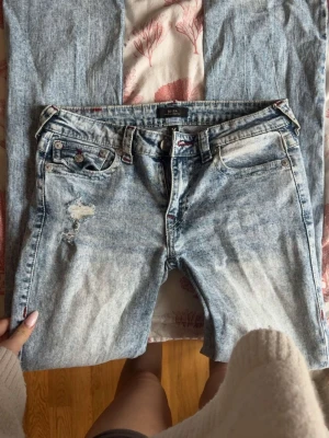 True Religion ljusblå slitna jeans - Säljer ett par ljusblå jeans från True Religion med stentvättad look och slitningar på vänster lår. Klassisk femficksmodell med dekorativa sömmar och logga på bakfickan. Perfekta för en avslappnad och trendig stil. Dem är omsydda till lågmidjat 