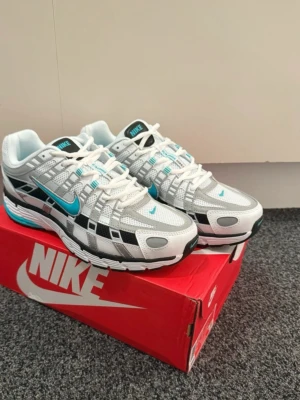 Nike vita och turkosa sneakers - Nike sneakers i vitt, grått och svart med turkosa detaljer. Skorna har en låg siluett, meshpaneler för andningsförmåga och klassisk Nike Swoosh på sidan. Snygg design med retrovibbar och robust yttersula. Perfekta för dig som gillar streetwear och sportig stil.