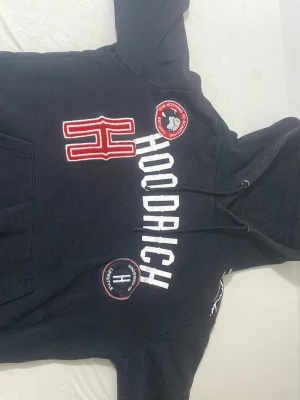 Svart Hoodrich hoodie med tryck - Snygg svart hoodie från Hoodrich med stora vita bokstäver på bröstet och röda detaljer. Märkets logga finns både som patch och tryck framtill. Klassisk huva med dragsko och känguruficka. Perfekt för en streetwear-look.