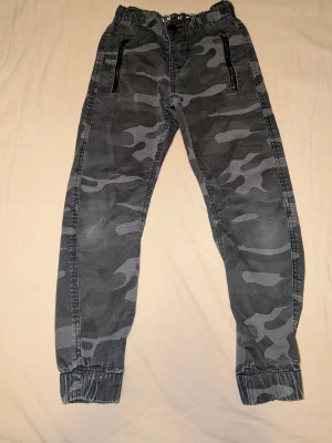 Stl 134 Camobyxor med dragkedjor👖 - Snygga byxor i grått camomönster med resår i midjan och benslut. Byxorna har två svarta dragkedjefickor framtill. Perfekta för en avslappnad och trendig stil. 