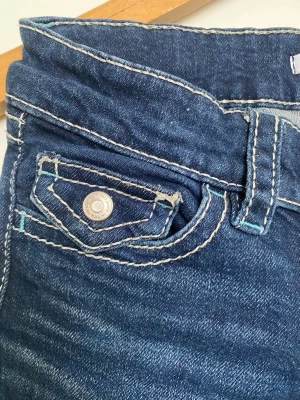 Mörkblå bootcut jeans med kontrastsömmar - Säljer ett par supersnygga mörkblå bootcut jeans med vita kontrastsömmar och snygga detaljer på fickorna. Jeansen har låg midja, knappstängning och klassisk femficksdesign. Materialet är denim strech och passformen är utsvängd nedtill.