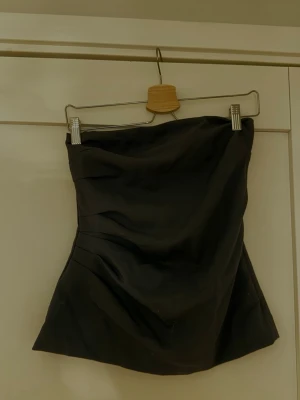 Svart bandeau topp från Lindex - Trendig svart bandeau topp från Lindex i storlek XS. Toppen har en figurnära passform och är sydd i ett slätt, glansigt material som ger en snygg siluett. Perfekt att styla med jeans eller kjol för en cool look.