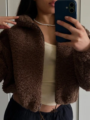 Brun croppad teddyjacka - Mysig brun teddyjacka i croppad modell med stora, fluffiga ärmar och bred krage. Jackan har en trendig oversized passform och är gjord i mjukt, lurvigt material som ger en cool och avslappnad look.