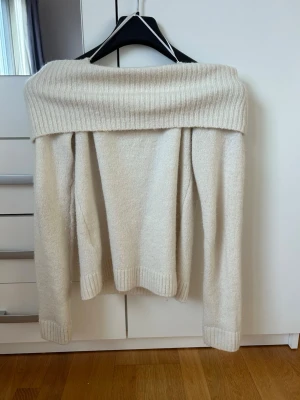 Off shoulder stickad tröja beige - Trendig off shoulder stickad tröja i beige med bred ribbad kant upptill och vid ärmslut samt nederkant. Mjuk och skön kvalitet, perfekt för lager på lager. Långärmad och rak passform.