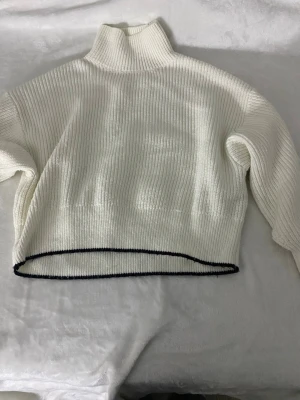 Vit stickad polotröja med svart kant - Vit stickad polotröja med ribbad struktur och svart kant längst ner. Tröjan har en hög krage och långa ärmar, perfekt för kyliga dagar. Oversized passform ger en avslappnad och trendig look.