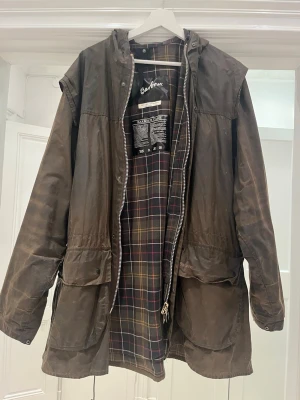 Brun vaxad parkas från Barbour - Klassisk brun vaxad parkas från Barbour med rutigt innerfoder i brunt, gult och rött. Jackan har huva, dragkedja och tryckknappar, samt stora fickor framtill. Perfekt för dig som gillar tidlös brittisk stil och funktionella ytterplagg.