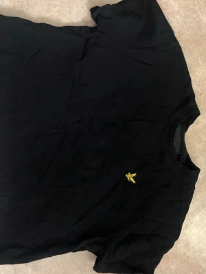 Svart t-shirt lyle and scott - Snygg svart t-shirt från Lyle & Scott med klassisk gul broderad logga på bröstet. T-shirten har rund hals och korta ärmar, perfekt till alla outfits. Tillverkad i mjuk bomull för skön känsla hela dagen.