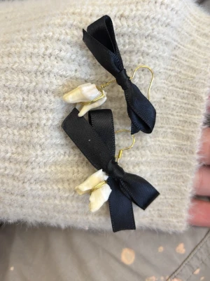 Örhängen med svarta rosetter och pärlor - Unika örhängen med svarta sidenrosetter och oregelbundna vita pärlor. Krokarna är i guldfärgad metall och designen är lekfull men ändå elegant. Perfekta för dig som vill sticka ut med accessoarerna.