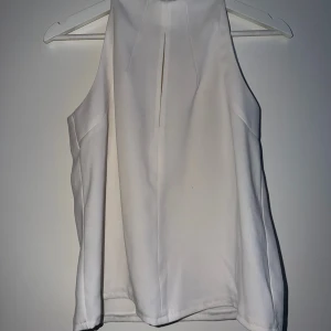Vit ärmlös halterneck topp - Snygg vit ärmlös topp med halterneck och stilren slitsdetalj framtill. Perfekt till jeans eller kjol för en clean look. Tillverkad i ett mjukt, lätt material som känns skönt mot huden.