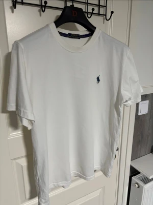 Vit t-shirt från Polo Ralph Lauren - Klassisk vit t-shirt från Polo Ralph Lauren med rund halsringning och den ikoniska blå logotypen broderad på bröstet. Tillverkad i mjuk bomull och har en normal passform. Perfekt basplagg för alla tillfällen.