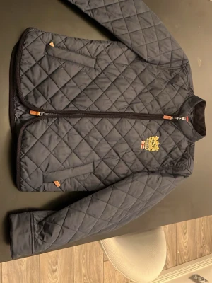 Marinblå quiltad vindjacka Morris - Snygg marinblå quiltad vindjacka från Morris med guldbroderad logga och brittisk flagga på bröstet. Jackan har två framfickor med dragkedja och detaljer i brunt läder. Klassisk ståkrage och smidig dragkedja framtill. Perfekt för dig som gillar stilren och sportig look. Säljer den för att jag behöver bli av med några jackor, absolut inget fel på den. Nypris 1400kr 
