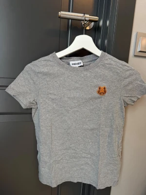 Grå t-shirt från Kenzo med tiger - Säljer en grå t-shirt från Kenzo med korta ärmar och rund hals. På bröstet finns ett broderat tigerhuvud i orange och svart som ger ett coolt statement. T-shirten är gjord i mjuk bomull och har en normal passform.