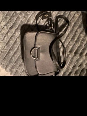 Svart axelväska  - Snygg svart axelväska i stilren design med lock och dekorativt spänne framtill. Väskan har justerbar axelrem och är tillverkad i ett slätt material som ger en modern look. Perfekt för att bära dina viktigaste grejer.