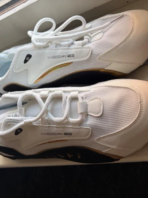 Vita sneakers  - Snygga vita sneakers med svarta och guldiga detaljer. Ovandelen är i mesh och syntet, med coola grafiska tryck på sidan. Skorna har en låg silhuett och vita snören. Perfekta för dig som gillar stilrena men ändå unika sneakers.