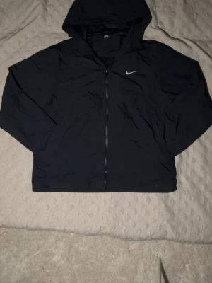 Svart Nike vindjacka med huva - Svart vindjacka från Nike med hel dragkedja och huva. Jackan har en enkel och sportig design med Nike-logga på bröstet. Tillverkad i lätt och vindtätt material, perfekt för blåsiga dagar. Klassisk passform och diskreta detaljer.