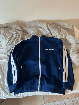 Blå Palm Angels hoodie med dragkedja - Snygg marinblå hoodie från Palm Angels med vita ränder längs ärmarna och vit dragkedja framtill. Loggan är tryckt på bröstet. Hoodien har två fickor med dragkedja och sportig design, perfekt för en streetwear-look.