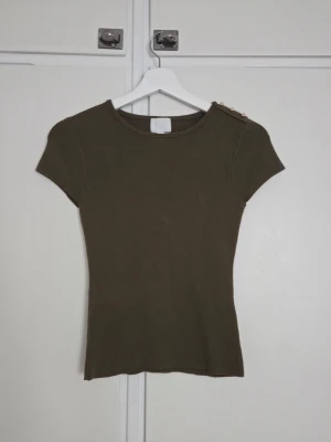 Olivgrön ribbad topp med guldfärgade knappar - Snygg olivgrön ribbad topp med korta ärmar och rund halsringning. Toppen har dekorativa guldfärgade knappar på ena axeln som ger en extra detalj. Perfekt att styla med jeans eller kjol för en trendig look.
