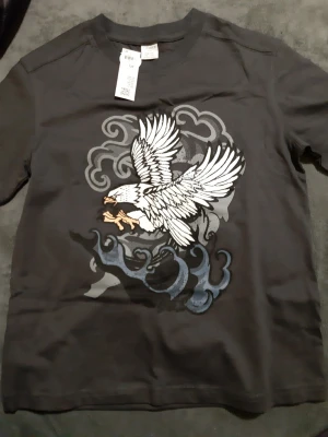 50 kr/ st. Mörkgrå t-shirt med örntryck 2 st. Helt nya. - Cool mörkgrå  t-shirt från Lindex, med ett stort tryck av en vit örn som flyger över moln i gråa och vita toner. T-shirten har rund hals och korta ärmar. Perfekt för dig som gillar grafiska motiv och streetstyle. Helt nya 2 st.