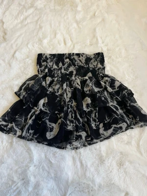 Svart kortkjol med volanger KappAhl - Snygg svart kortkjol från KappAhl med vitt abstrakt mönster och bred smock i midjan. Kjolen har flera lager volanger som ger en luftig och trendig look. Perfekt för dig som gillar att sticka ut med coola detaljer.