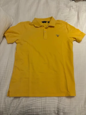 Gul piképolo från Gant - Snygg gul piképolo från Gant med klassisk krage och knappar framtill. Diskret Gant-logga med broderad sköld på bröstet. Perfekt till jeans eller shorts för en fräsch och sportig look. Hör av er vid mer info storlek 158-164 13-14 years old!
