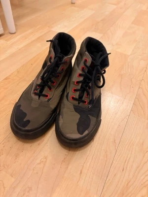 Kamouflagefärgade high top sneakers från Ralph lauren - Säljer ett par high top sneakers med kamouflagemönster i grönt, svart och beige. Skorna har svarta sulor, svarta skosnören och röda öljetter som ger en cool kontrast. Perfekta för dig som vill sticka ut med din streetwear-stil. Dem är äkta Ralph lauren