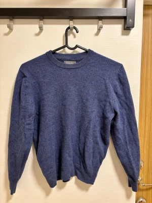 Mörkblå stickad tröja i merinoull  - En enkel och stilren mörkblå stickad tröja med rund halsringning och långa ärmar. Tröjan har ribbade muddar vid hals, ärmslut och nederkant. Perfekt att matcha med jeans eller kjol för en avslappnad look.