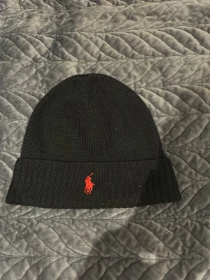Svart mössa från Polo Ralph Lauren - Svart ribbstickad mössa från Polo Ralph Lauren med klassisk röd logobrodyr framtill. Mössan har uppvikt kant och är tillverkad i mjukt material som håller dig varm. Perfekt för dig som vill ha en stilren och ikonisk accessoar.