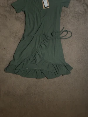 Grön omlottklänning med volang - Snygg grön kortklänning i omlottmodell med volangdetaljer längs nederkanten och knytband i midjan. Perfekt för dig som gillar en enkel men trendig stil. Kortärmad och i mjukt material som känns skönt mot huden.