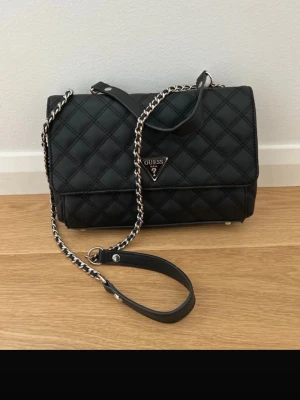 Svart quiltad handväska från Guess - Snygg svart quiltad axelväska från GUESS med kedjerem och justerbart axelband.  Elegant och tidlös design som passar både till vardag och fest.  Logotyp i metall framtill  Färg: Svart  Material: Syntetiskt läder  Nypris i butik: 2,600 kr  Väskan är i fint skick och sparsamt använd. Observera att färgen på kedjan är något sliten, men detta påverkar inte funktionen och knappt helhetsintrycket. Perfekt för dig som vill ha en lyxig men praktisk väska som lyfter varje outfit!